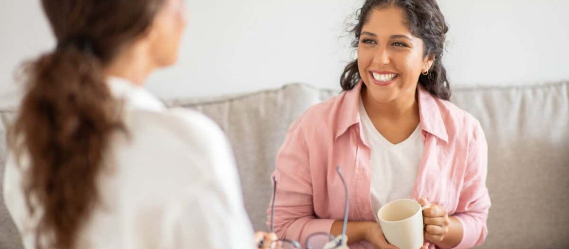 A woman wearing a pink shirt sits on a couch and smiles while holding a cup of coffee. She is talking to another woman sitting next to her, who is wearing a white shirt.
