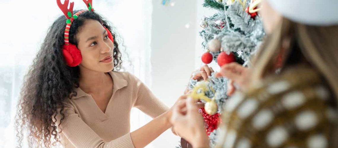 African American woman decorate a Christmas tree with ornaments and lights during Christmas eve celebration. The woman's vibrant energy shines through carefully places ornaments on the Christmas tree. Earmuffs add a touch of warmth and style to her festive ensemble.