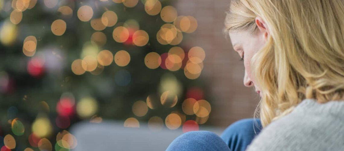 A Caucasian woman is indoors in her living room. There is a Christmas tree in the background. The woman is wearing warm clothing. She is sitting on the couch and looking sad because she is alone on Christmas day.