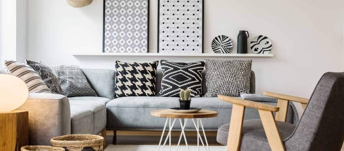 small table standing between a sofa with pillows and an armchair on a white rug in boho living room interior with posters on a shelf