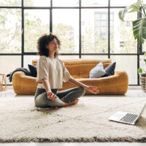 Young multiracial latina woman meditating at home with online video meditation lesson using laptop.