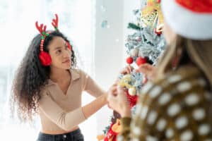 African American woman decorate a Christmas tree with ornaments and lights during Christmas eve celebration. The woman's vibrant energy shines through carefully places ornaments on the Christmas tree. Earmuffs add a touch of warmth and style to her festive ensemble.