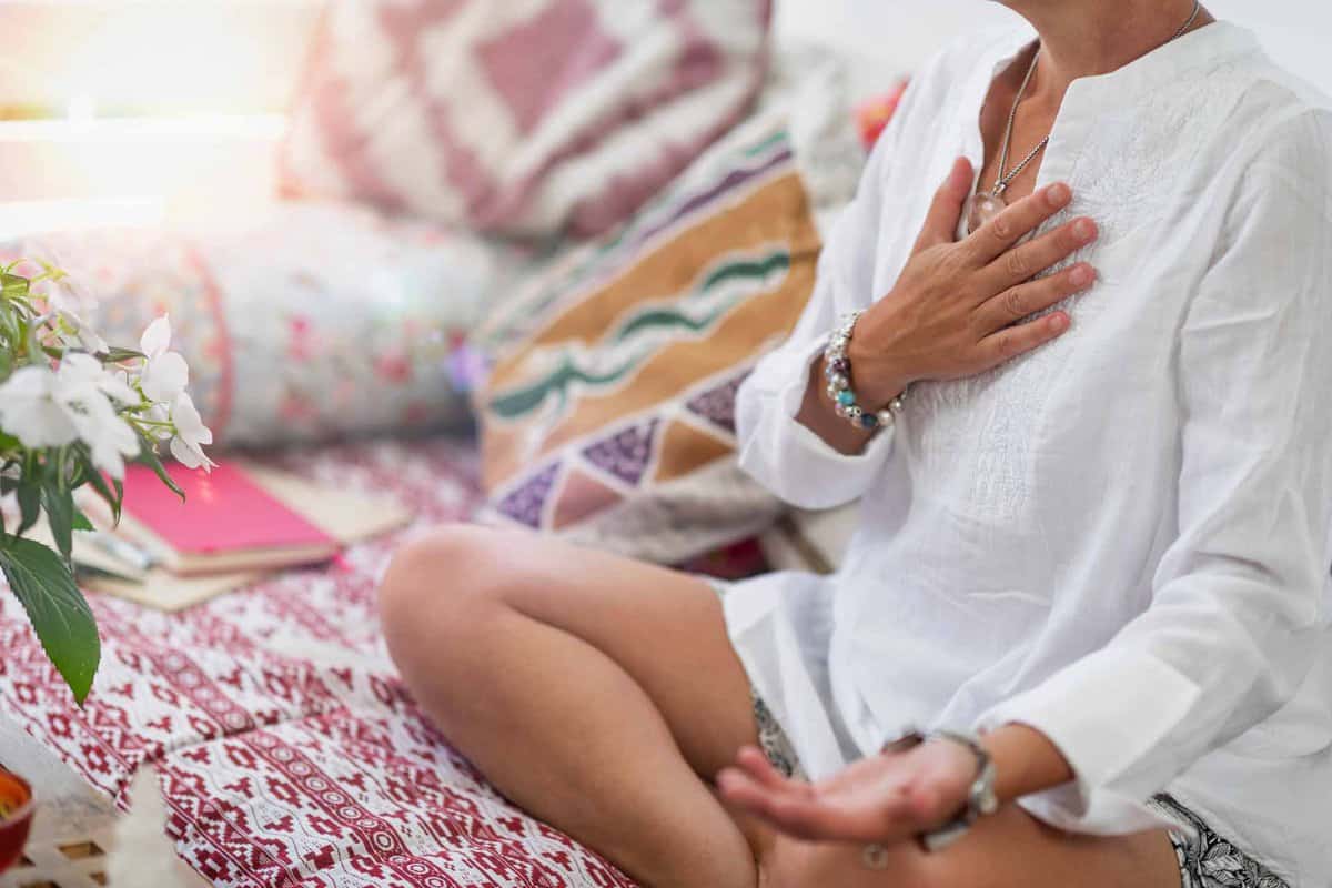 Self-Healing Heart Chakra Meditation. Woman sitting in a lotus position with right hand on heart chakra and left palm open in a receiving gesture. Self-Care Practice at Home